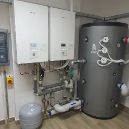 Dwie białe jednostki wewnętrzne pompy ciepła Panasonic Aquarea zamontowane obok szarego zbiornika akumulacyjnego ciepła, widoczne liczne rury z zaworami i manometrami, na ścianie skrzynka...