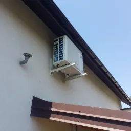 Zewnętrzna jednostka klimatyzacji zamontowana na elewacji budynku z widoczną rurą odprowadzającą skropliny i brązowym orynnowaniem.