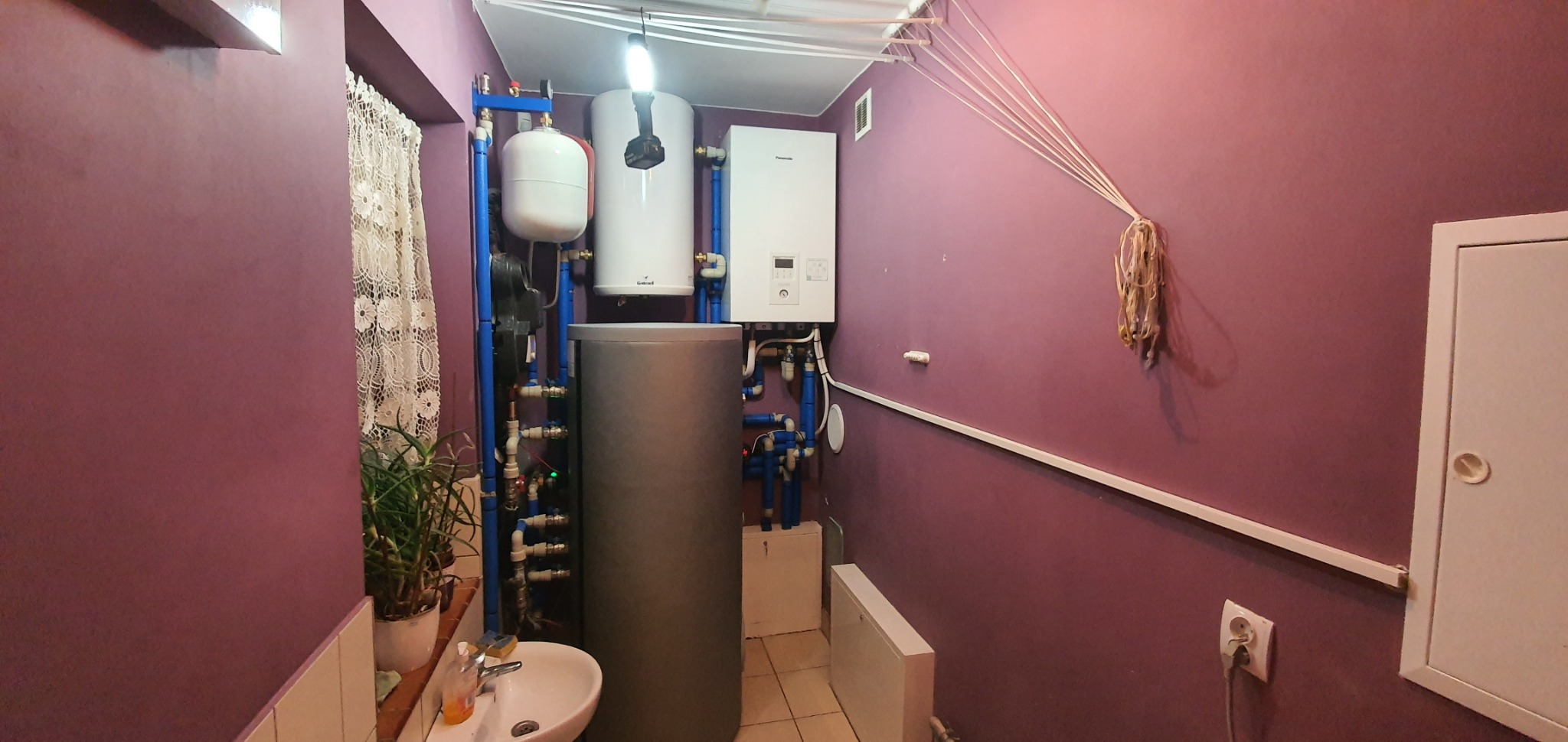 Kotłownia z dwoma zbiornikami, białym i szarym, połączonymi niebieskimi rurami, na tle fioletowej ściany. Dodatkowo widoczny biały bojler, umywalka z rośliną i suszarka na pranie pod sufitem.
