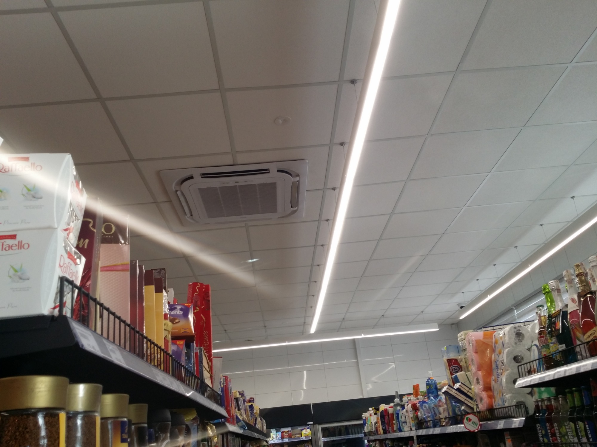 Kasetonowy klimatyzator sufitowy w supermarkecie, widoczny na tle oświetlenia liniowego i półek z towarem.