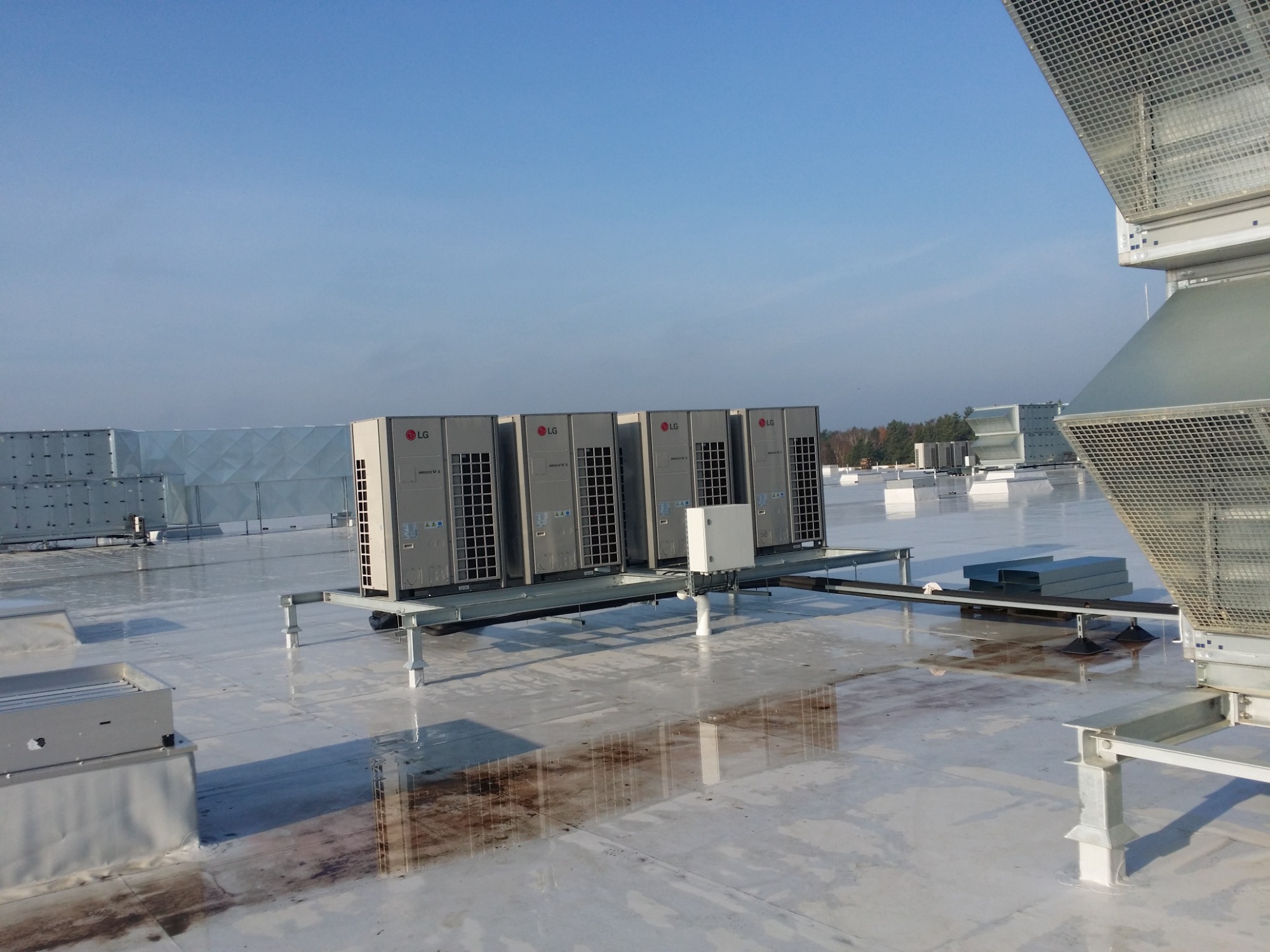 Rząd jednostek zewnętrznych klimatyzacji LG Multi V 5 zamontowanych na dachu budynku, na metalowej konstrukcji wsporczej, widoczne odbicia w kałużach na dachu.