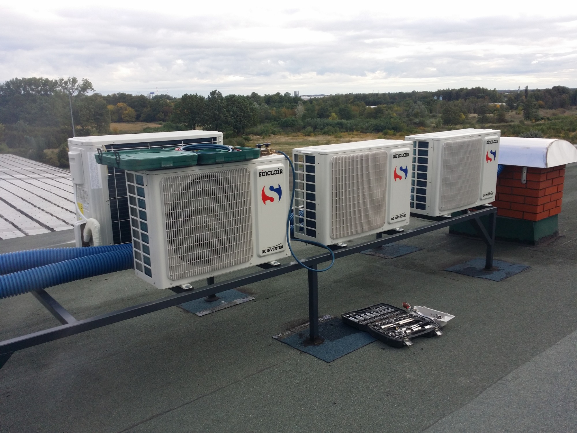 Rząd białych jednostek zewnętrznych klimatyzacji Sinclair DC Inverter na dachu, z skrzynką narzędziową obok, na tle zieleni i pochmurnego nieba.
