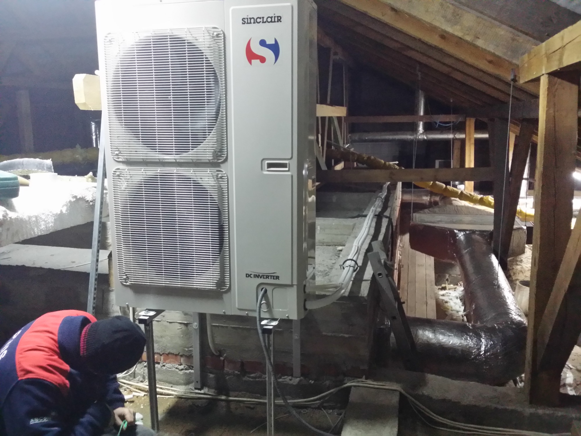 Instalacja klimatyzatora Sinclair DC Inverter w surowym strychu z widoczną konstrukcją drewnianą dachu, pracownik w czerwono-niebieskiej kurtce podłącza przewody.