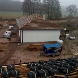 Montaż brązowej dachówki na drewnianej konstrukcji dachu jednopiętrowego domu w trakcie budowy, widoczny przyczepka firmy dekarskiej Cichosz i stosy czarnej dachówki na pierwszym planie, mglista...