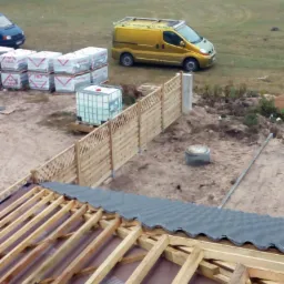 Konstrukcja drewniana dachu z częściowo ułożonymi szarymi dachówkami, widok z góry na budowę, w tle żółty samochód dostawczy i palety z materiałami budowlanymi.