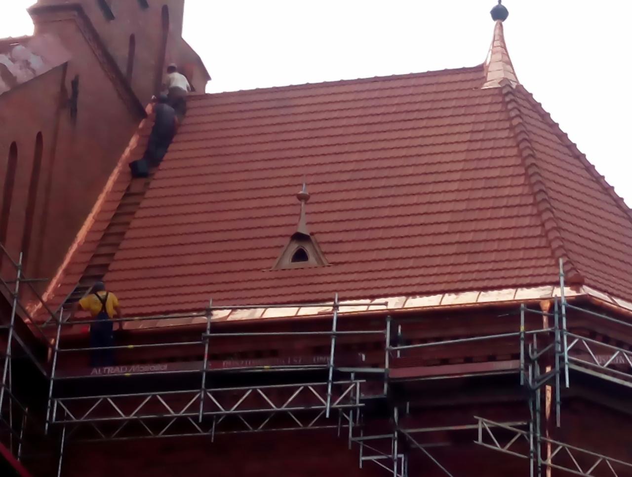 Czerwona dachówka na kościelnej wieży podczas montażu, dekarze na rusztowaniu i na dachu, widoczny detal architektoniczny w postaci małej wieżyczki z oknem.