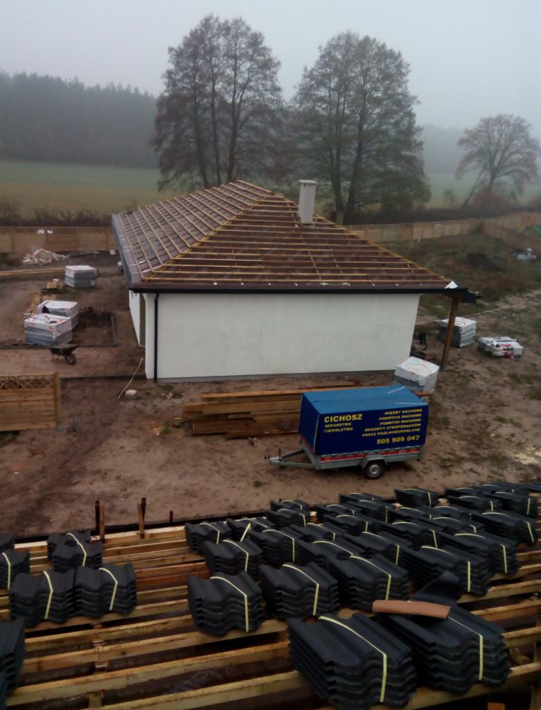 Montaż brązowej dachówki na drewnianej konstrukcji dachu jednopiętrowego domu w trakcie budowy, widoczny przyczepka firmy dekarskiej Cichosz i stosy czarnej dachówki na pierwszym planie, mglista...