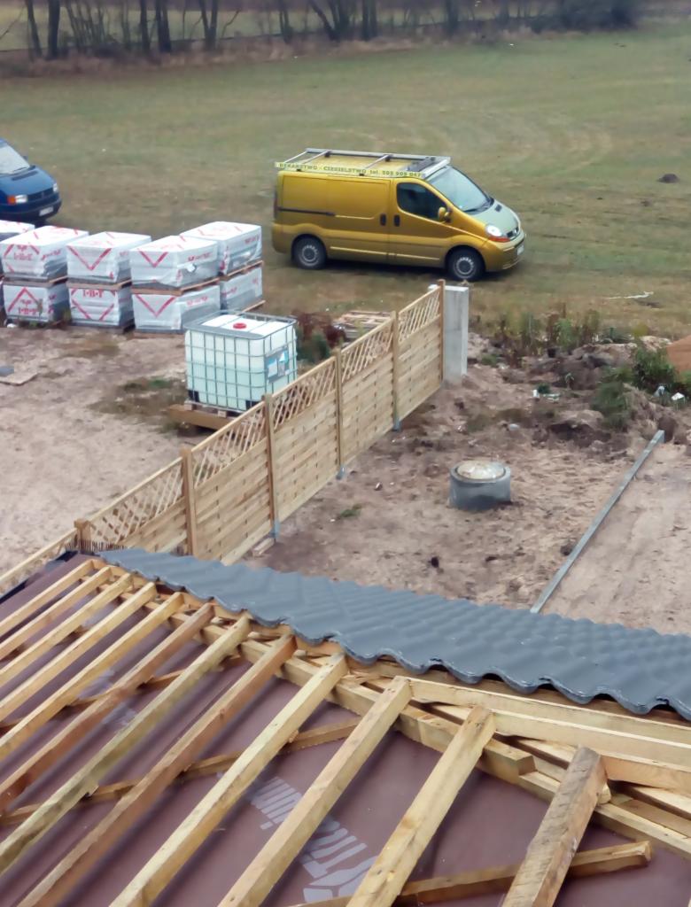 Konstrukcja drewniana dachu z częściowo ułożonymi szarymi dachówkami, widok z góry na budowę, w tle żółty samochód dostawczy i palety z materiałami budowlanymi.