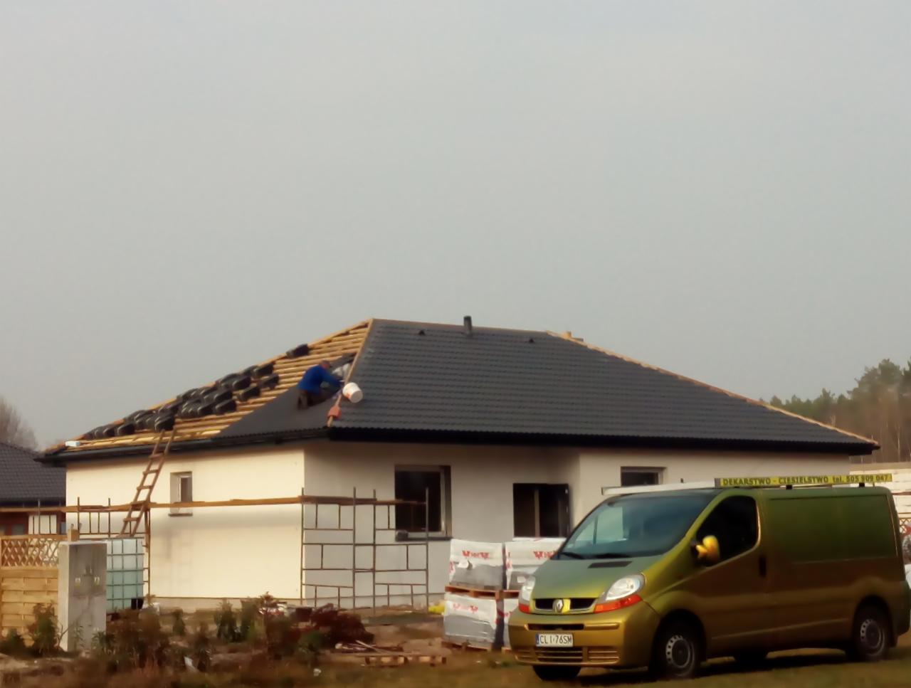 Prace na dachu domu jednorodzinnego: dekarz w niebieskiej kurtce układa czarne dachówki, widoczna drabina, rusztowanie, zaparkowany zielony bus firmy dekarskiej z reklamą na boku.