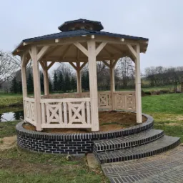 Drewniana altana ogrodowa z czarnym dachem, otoczona murkiem z cegły i schodami z kostki, w tle widoczny staw i drzewa.