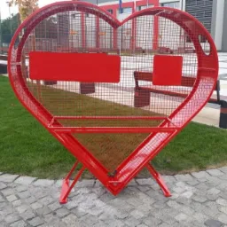 Czerwona, metalowa konstrukcja w kształcie serca z siatki, ustawiona na zewnątrz na kostce brukowej, z prostokątnymi elementami do mocowania napisów lub zdjęć.