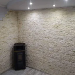 Ściana wyłożona dekoracyjną, piaskową okładziną imitującą kamień, obok stoi czarny piec wolnostojący, sufit podwieszany z punktowym oświetleniem.