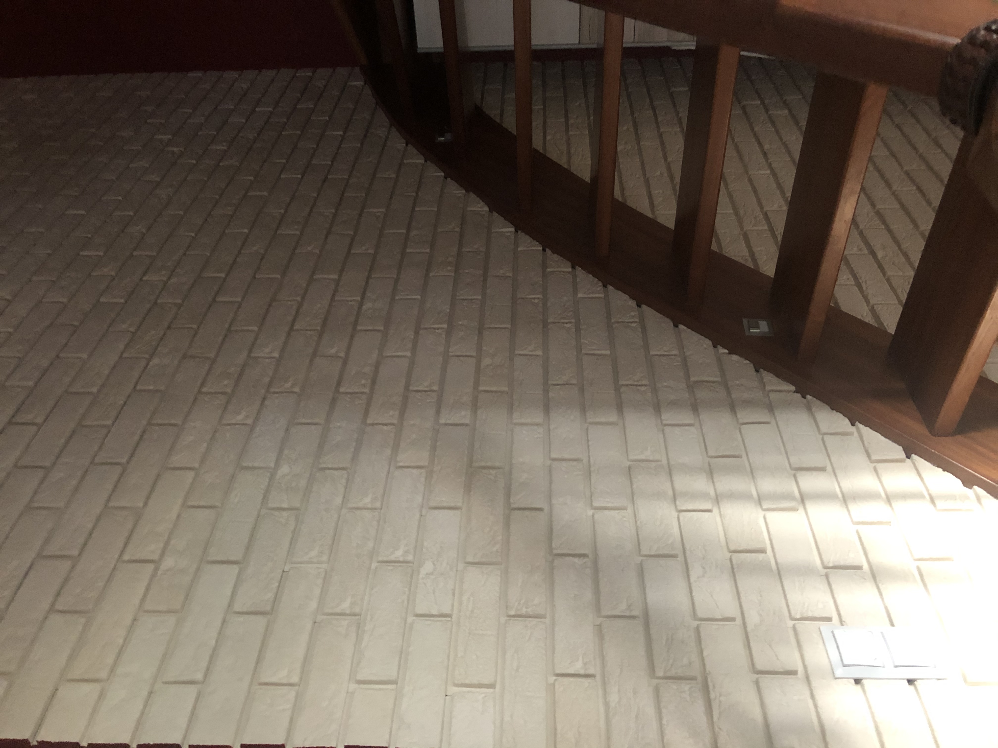 Wykończona ściana z białych, podłużnych płytek imitujących cegłę, ułożonych pod schodami z drewnianą balustradą, z widocznym gniazdkiem elektrycznym.