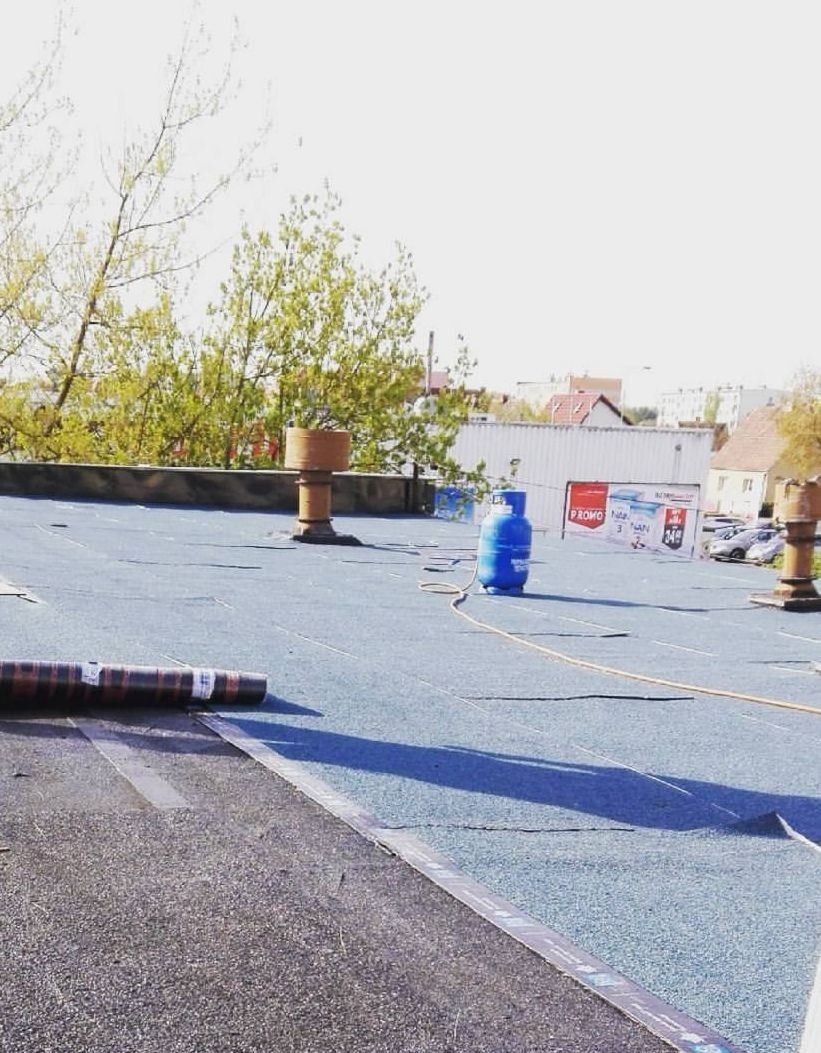 Niebieski dach pokryty papą z widocznym cylindrem wentylacyjnym, butlą gazową i rolką papy w trakcie prac dekarskich, z budynkami w tle.