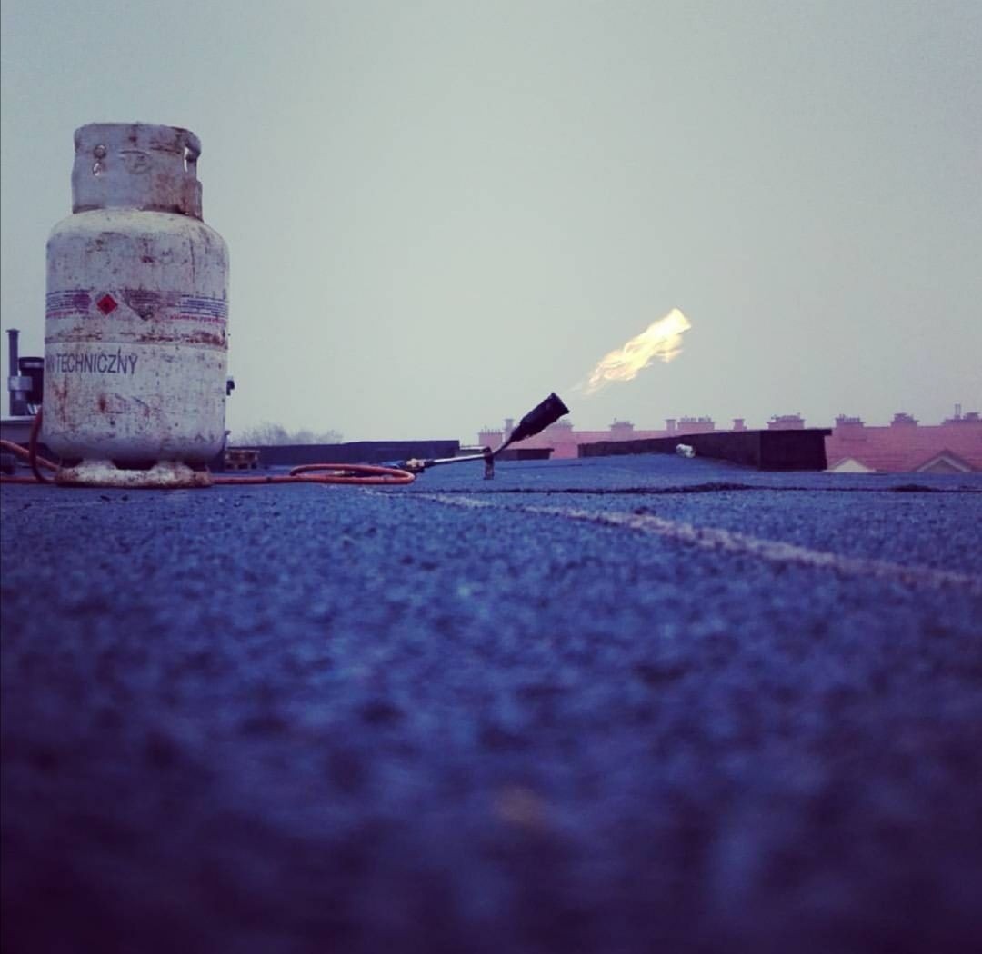 Butla gazowa i palnik dekarski podczas prac na pokrytym papą dachu, z widocznym płomieniem i budynkami w tle.