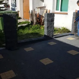 Nowo ułożona, czarno-żółta kostka brukowa przed domem z dwoma kamiennymi słupkami, częściowo widoczna postać zamiatająca bruk.