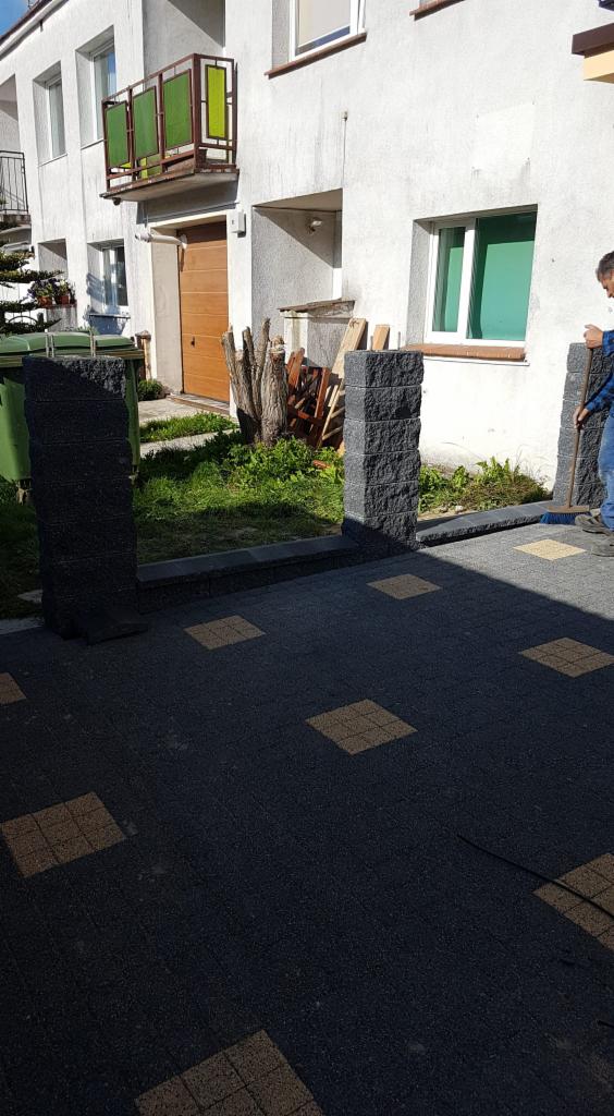 Nowo ułożona, czarno-żółta kostka brukowa przed domem z dwoma kamiennymi słupkami, częściowo widoczna postać zamiatająca bruk.