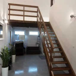 Wnętrze z drewnianymi schodami prowadzącymi na antresolę, białe ściany, szara podłoga, widoczne rośliny doniczkowe i meble w tle, surowe belki stropowe.