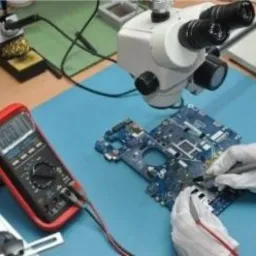 Płyta główna laptopa pod mikroskopem, diagnozowana multimetrem przez osobę w rękawiczkach ochronnych. Na biurku widoczna stacja lutownicza i inne narzędzia.