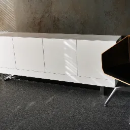 Nowoczesna biała komoda na metalowych nóżkach i designerskie krzesło z czarnym siedziskiem w minimalistycznym wnętrzu, oświetlonym promieniami słońca wpadającymi przez żaluzje.