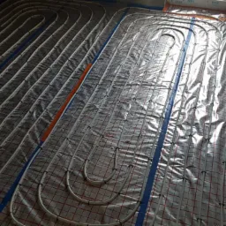 Białe rury ogrzewania podłogowego ułożone na srebrnej folii z czerwoną kratką, zabezpieczone niebieską taśmą, widoczne okna w tle.