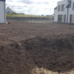 Przygotowany teren pod budowę z hałdami ziemi, widoczne fundamenty ogrodzenia i dwa budynki w tle.