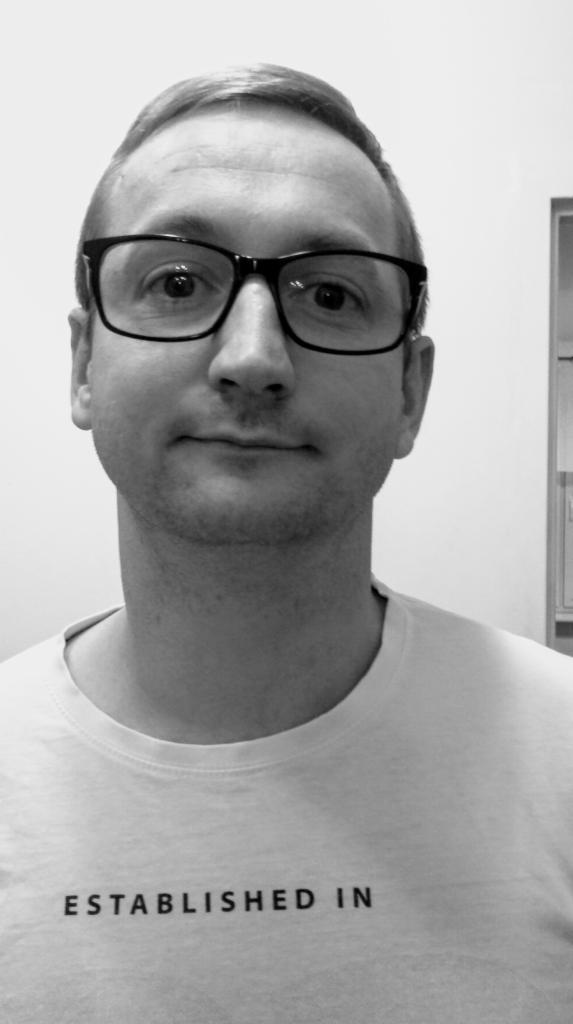 Czarno-białe zbliżenie portretowe mężczyzny w okularach i koszulce z napisem 'ESTABLISHED IN', na neutralnym tle.