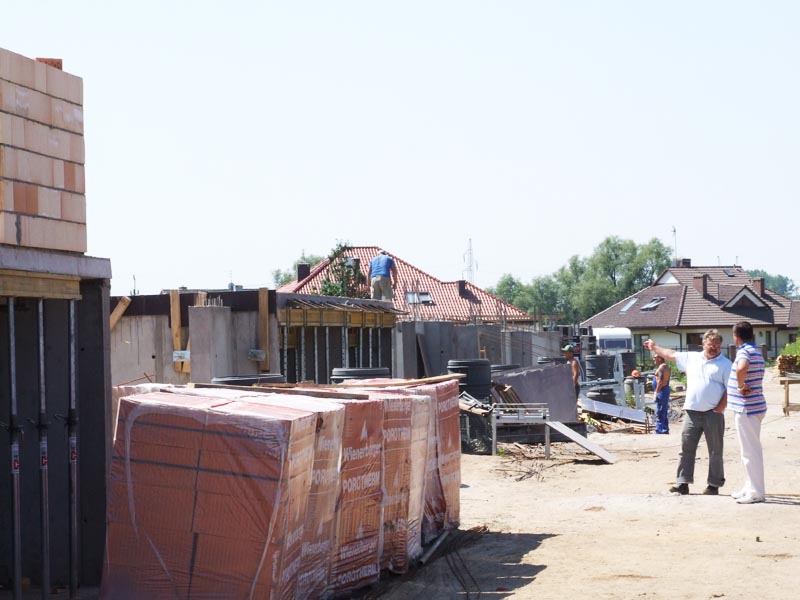 Budowa domu w trakcie realizacji: widoczne fundamenty, ściany z pustaków, przygotowana konstrukcja dachu, palety z materiałami budowlanymi, pracownicy na budowie i dwóch mężczyzn omawiających...