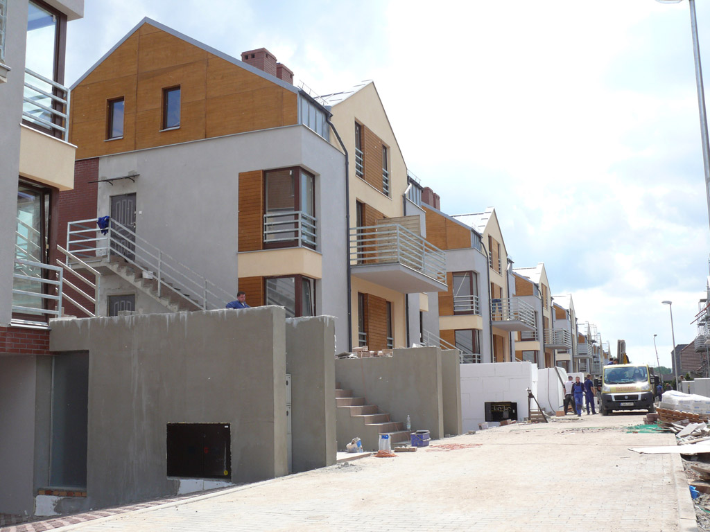 Szereg nowo wybudowanych domów w trakcie wykończenia, z widocznymi pracownikami i samochodem dostawczym na brukowanej drodze dojazdowej.