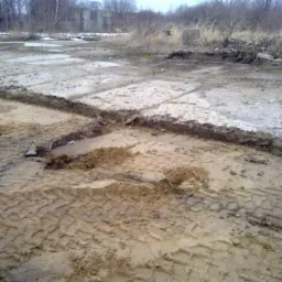 Używane, betonowe elementy nawierzchni z widocznymi śladami opon w błotnistym terenie. Część elementów jest uszkodzona, a teren wokół jest zaniedbany.