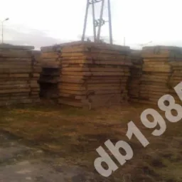 Stosy używanych, prostokątnych elementów betonowych składowane na nierównym terenie porośniętym trawą, widoczne słupy energetyczne w tle.