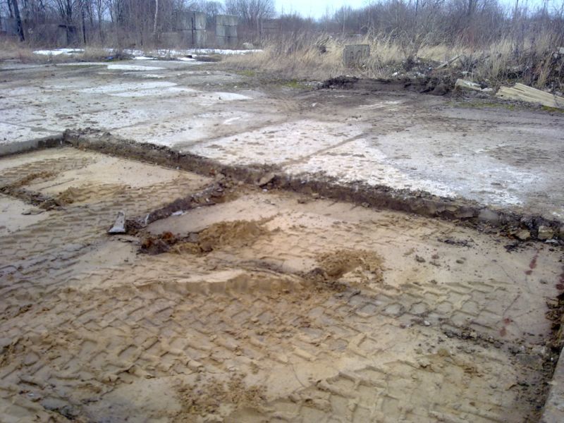 Używane, betonowe elementy nawierzchni z widocznymi śladami opon w błotnistym terenie. Część elementów jest uszkodzona, a teren wokół jest zaniedbany.