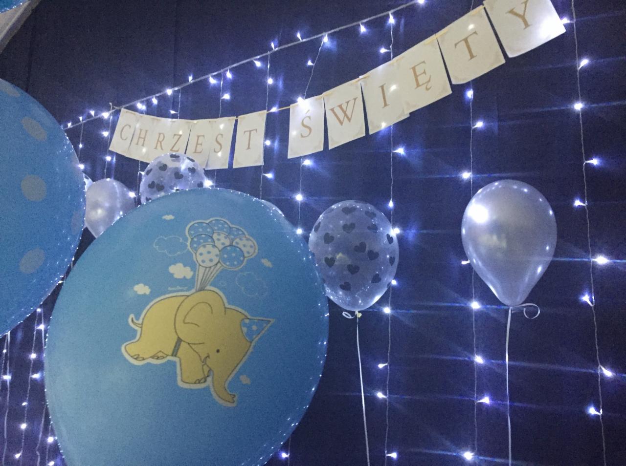 Dekoracja na chrzest: niebieskie balony z motywem słonia i balonów, transparent 'Chrzest Święty' na tle światełek.