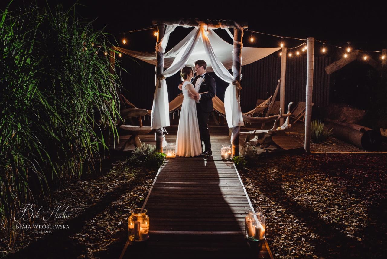 Para młoda w objęciach pod drewnianą altaną z białymi tkaninami i girlandami świetlnymi, wieczorny plener z lampionami na drewnianym podeście.