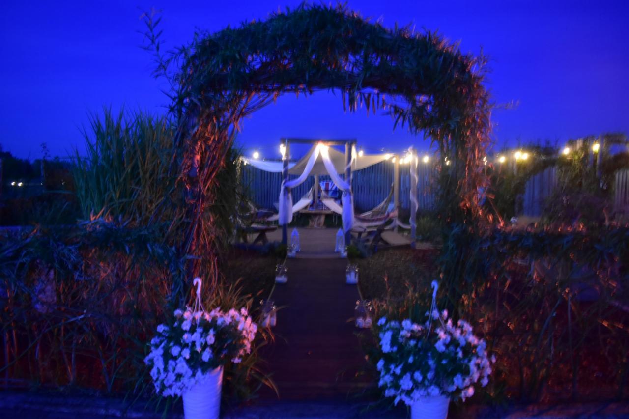 Uroczyste miejsce na zewnątrz z drewnianą ścieżką, łukiem z zieleni, białymi kwiatami w wiszących doniczkach i baldachimem z białych tkanin, oświetlone lampkami i niebieskim światłem.