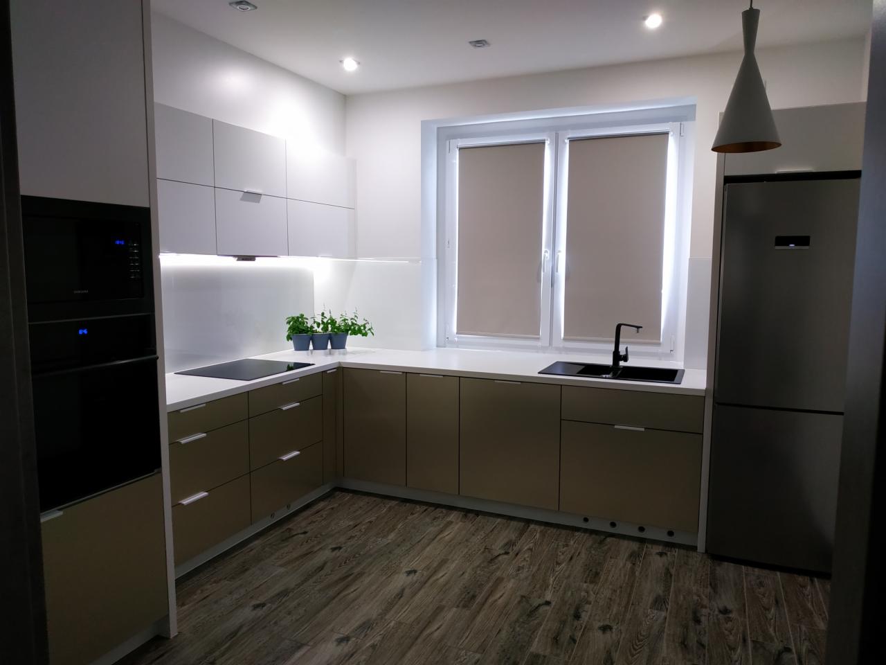 Nowoczesna kuchnia z beżowymi i białymi szafkami, drewnianą podłogą, czarnym zlewem i baterią oraz roletami w oknie, oświetlona lampą wiszącą.