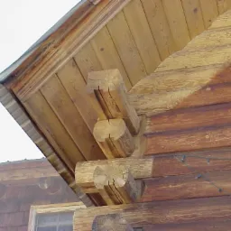 Fragment drewnianej elewacji domu z bali, z widocznymi wystającymi belkami stropowymi i fragmentem dachu. Na elewacji widoczne ślady po czyszczeniu, z jaśniejszym fragmentem po prawej stronie.