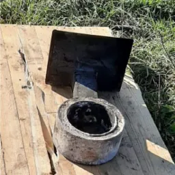 Metalowy element konstrukcyjny w kształcie kwadratu i okrągły element betonowy, oba po obróbce, leżą na drewnianej palecie na trawie.