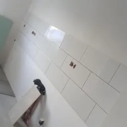 Ściana w trakcie wykańczania z białymi, prostokątnymi płytkami, widoczne otwory na gniazdka, na podłodze narzędzia i resztki materiałów budowlanych.