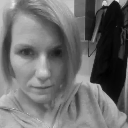 Portret kobiety w szarej bluzie z kapturem, z łazienką w tle i wiszącymi ubraniami na wieszaku.