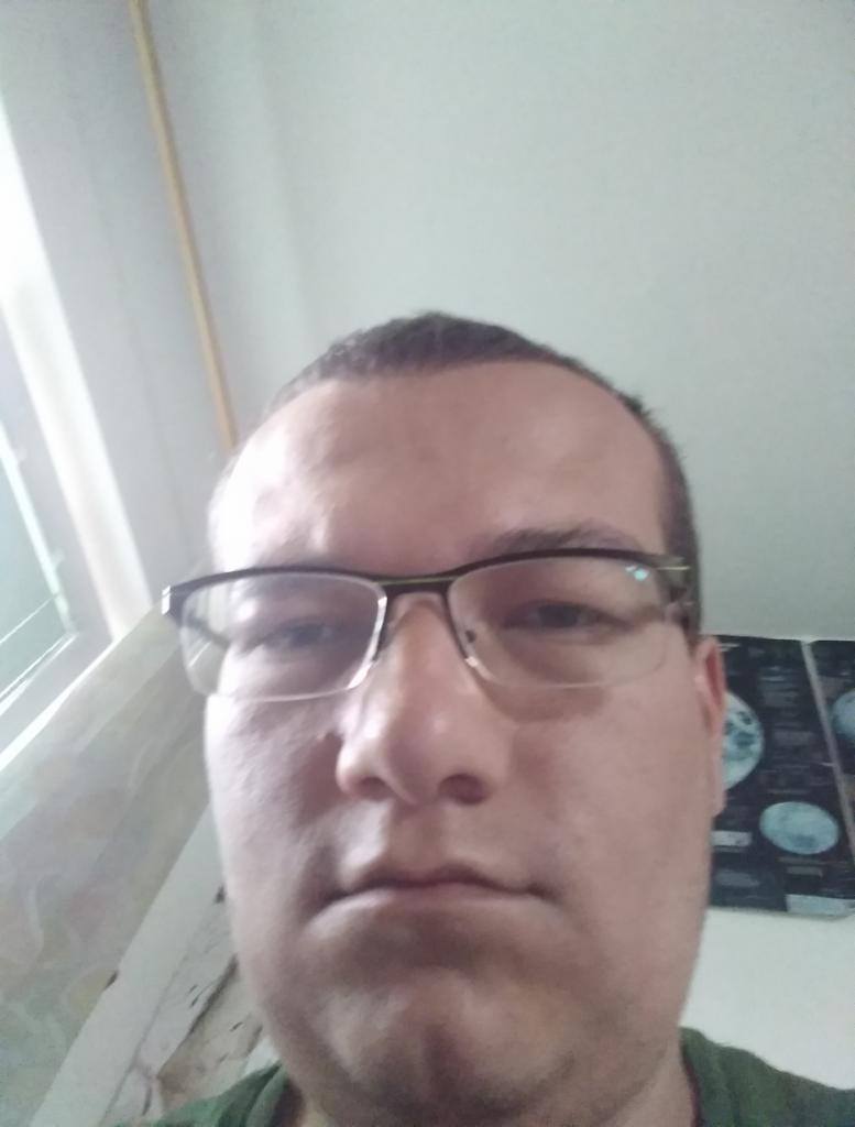 Autoportret mężczyzny w okularach z bliska, z widocznym oknem i plakatem z motywem kosmicznym w tle. Ujęcie z góry.