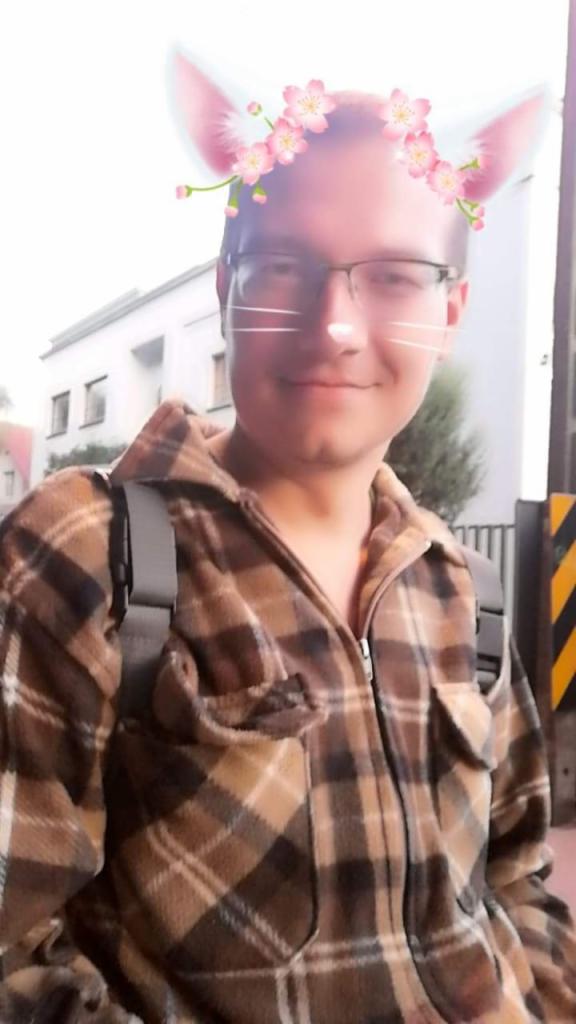 Młody mężczyzna w okularach i flanelowej kurtce pozuje do selfie z filtrem uszu kota i kwiatów. W tle budynek i fragment żółto-czarnej bariery.