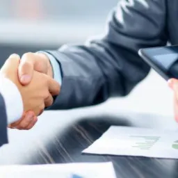 Uścisk dłoni nad stołem z dokumentami i tabletem. Biznesowe spotkanie, finalizacja umowy. Mężczyźni w garniturach. Stonowana kolorystyka, profesjonalny klimat.