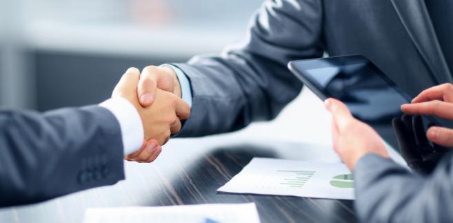 Uścisk dłoni nad stołem z dokumentami i tabletem. Biznesowe spotkanie, finalizacja umowy. Mężczyźni w garniturach. Stonowana kolorystyka, profesjonalny klimat.