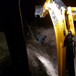 Pracownik w wykopie przy żółtej koparce JCB w słabym oświetleniu, prawdopodobnie przygotowuje dno wykopu pod rury.