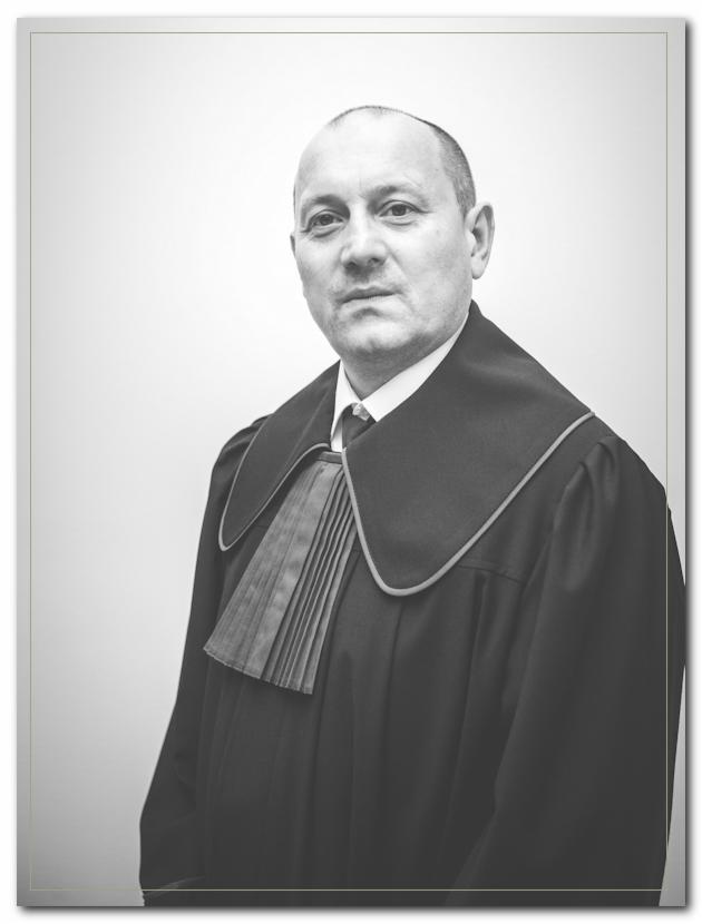 Portret mężczyzny w todze adwokackiej na jasnym tle, z delikatnym uśmiechem i widoczną fakturą materiału togi.