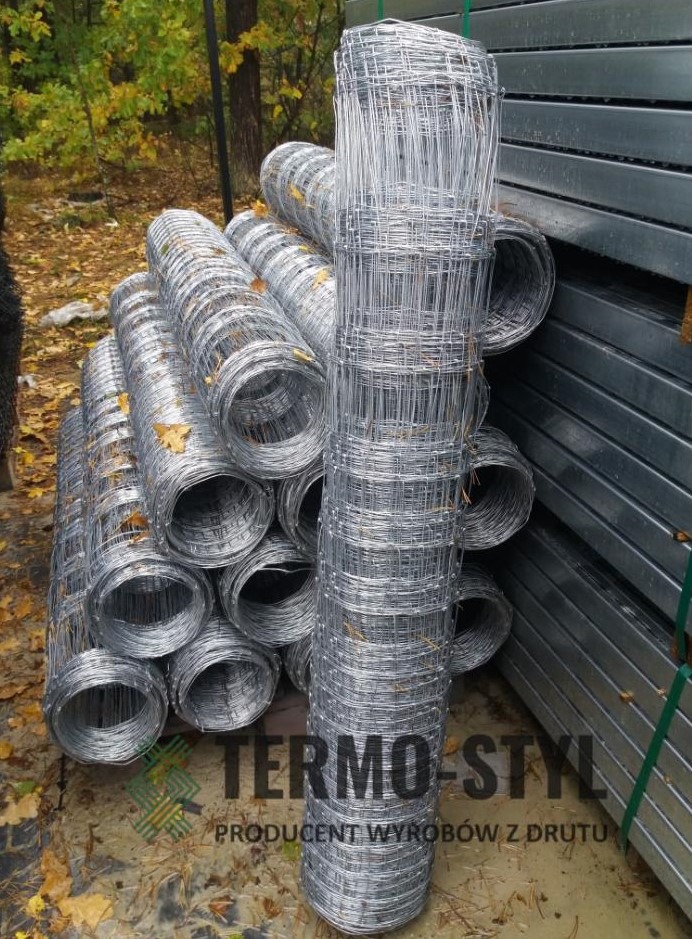 Stos rolek siatki ogrodzeniowej z drutu ocynkowanego, ułożonych na palecie, z widocznym logo producenta na pierwszym planie i jesiennym tłem.