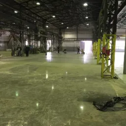 Wnętrze hali przemysłowej z betonową posadzką w trakcie czyszczenia. Widoczne metalowe konstrukcje nośne, żółte słupy, pracownicy z maszynami czyszczącymi oraz odblaski światła na powierzchni...