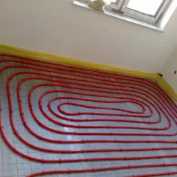 Czerwone rury ogrzewania podłogowego ułożone spiralnie na siatce, zabezpieczone żółtą folią przy ścianach w jasnym pomieszczeniu z oknem dachowym.