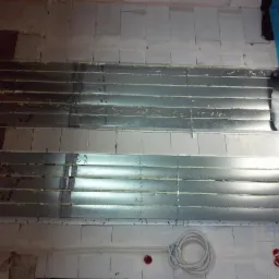 Montaż kanałów wentylacyjnych pod sufitem, widoczne blaszane rury z napisami, białe ściany z bloczków i przewody elektryczne.